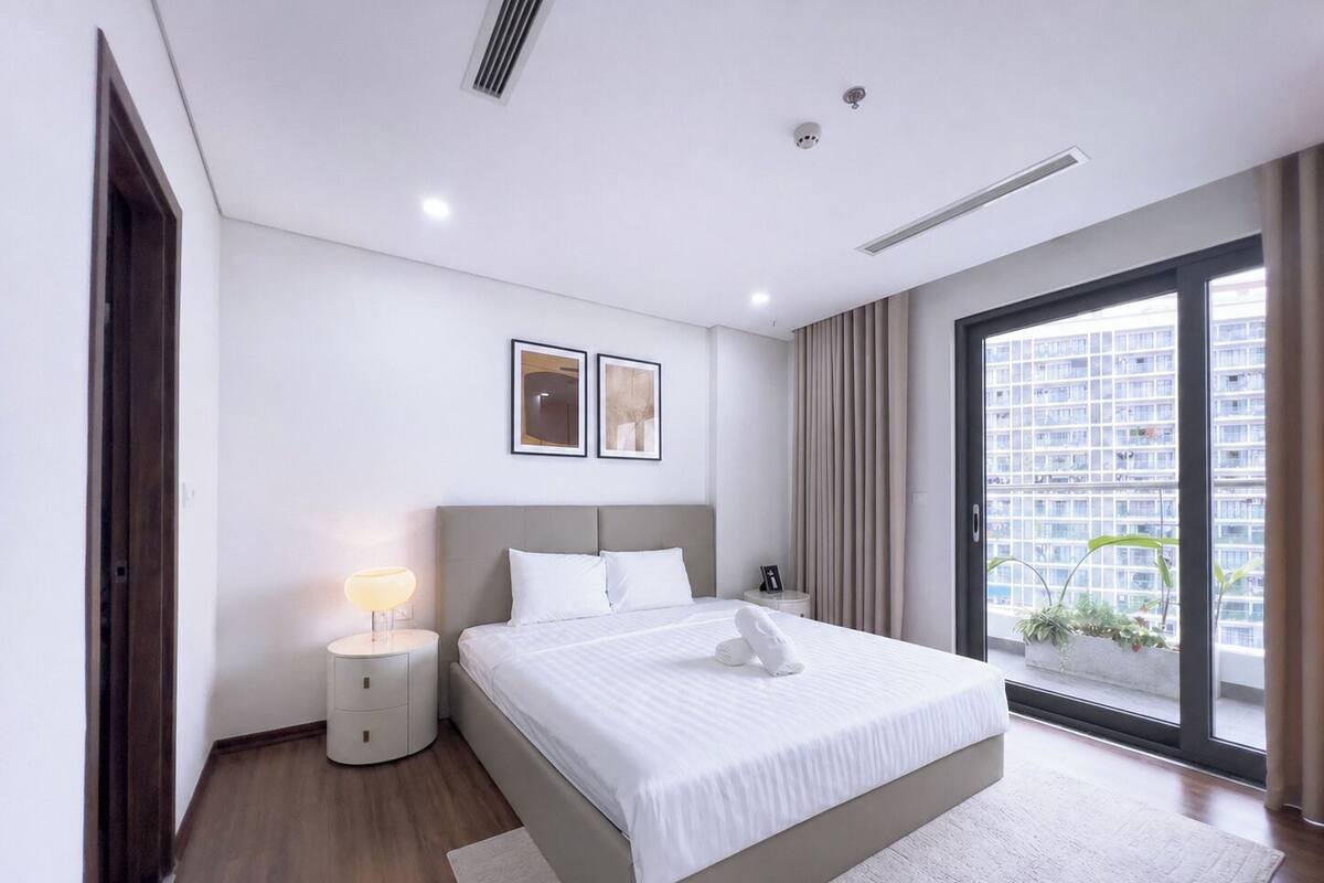 Quarto de hotel moderno com uma cama grande, mesas de cabeceira, uma janela com vista para edifícios e iluminação embutida no teto.