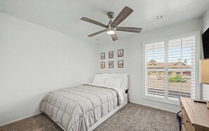 Ein Schlafzimmer mit Bett, Deckenventilator, Fenster mit Jalousien und Schreibtisch mit Lampe.