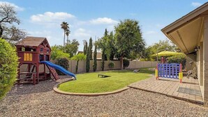 A backyard with a playhouse, a slide, a small lawn, and a patio area with a table and chairs. - Resort backyard and pool - close to everything (Mesa)