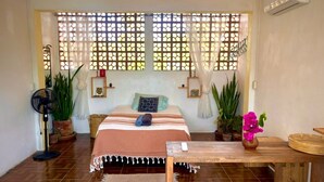 Een kamer met een bed, een ventilator, planten in potten en een raam met een lattenroede.