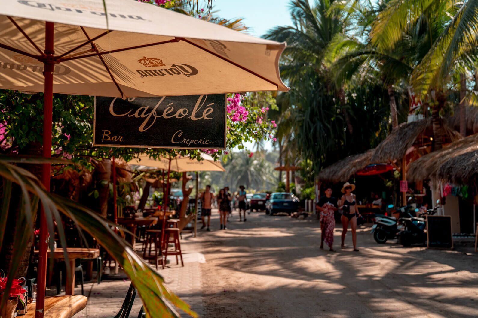 A café with outdoor seating, palm trees, and a thatched-roof structure.