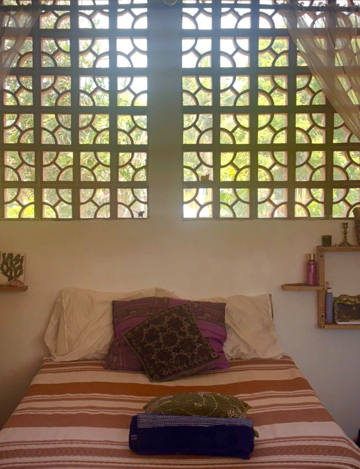 Ein Bett mit einem gestreiften Überwurf, zwei Kissen und einer gefalteten Decke. Ein Fenster mit geometrischem Muster und einem Vorhang ist zu sehen.