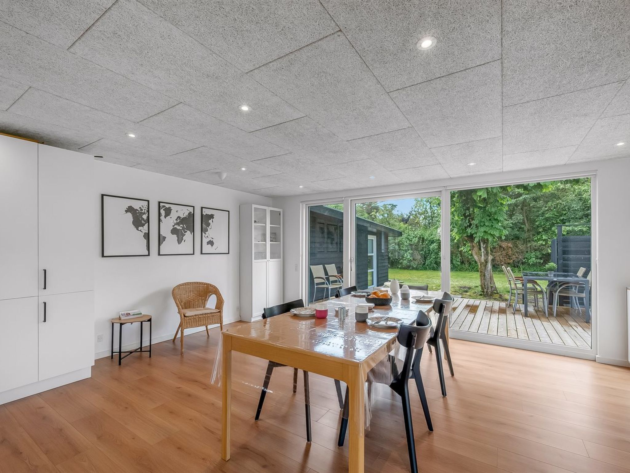 A spacious room with a wooden dining table, chairs, a wicker chair, a small side table, and a bookshelf. There is a large window providing a view of the outdoors.