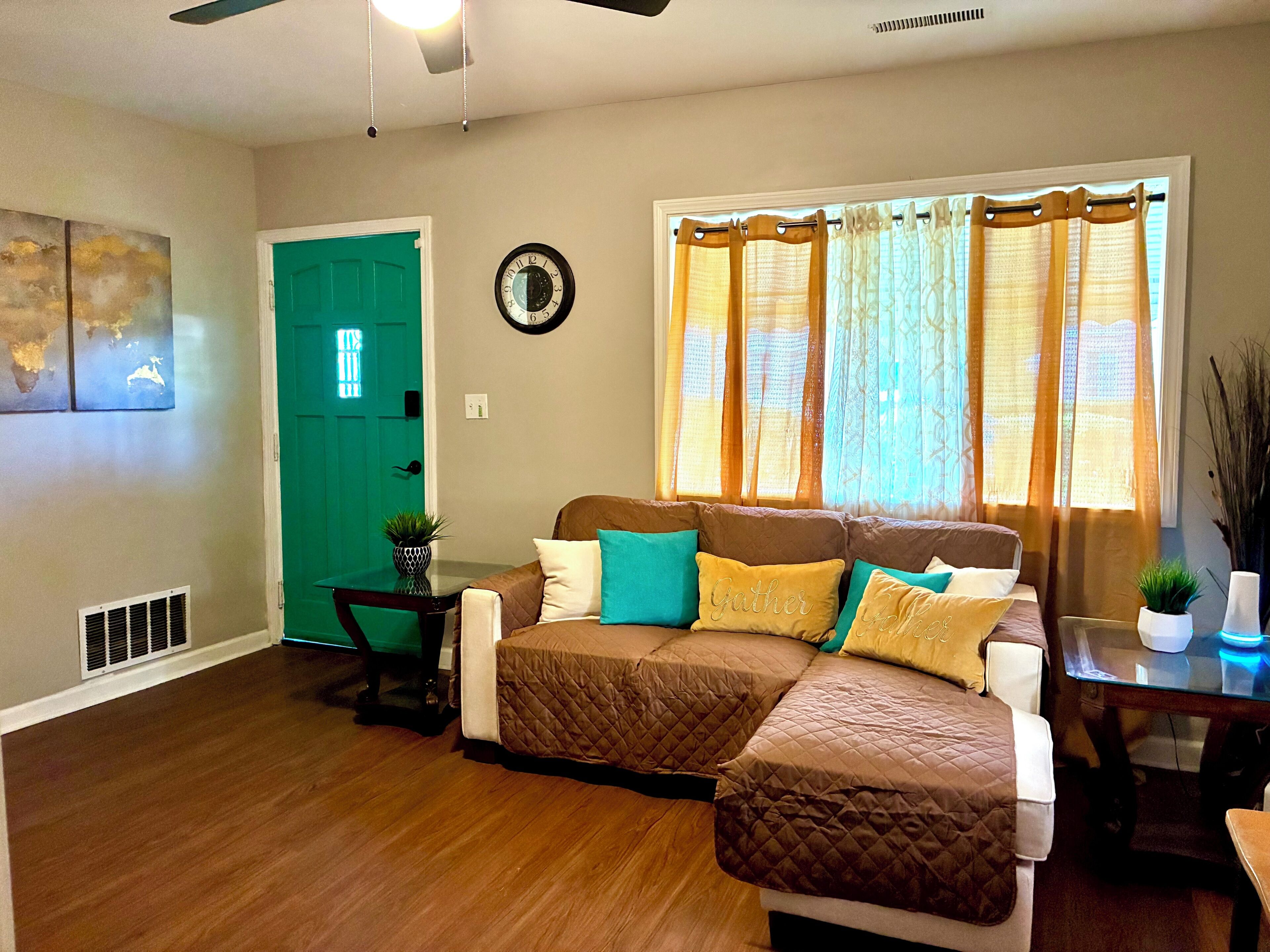 A living room with a sectional sofa, a green door, a wall clock, and a window with curtains.