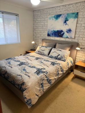 A bedroom with a bed, a nightstand, a lamp, a window, and a wall with a textured pattern. - NR 4 Cove Apartment Carrickalinga (Carrickalinga)