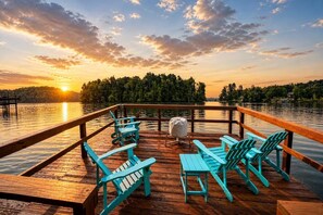 A wooden deck with turquoise Adirondack chairs and a grill, overlooking a lake with a sunset.