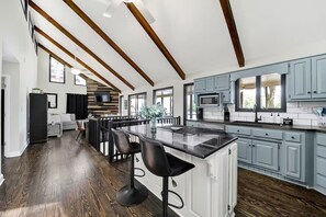 A modern kitchen with a central island, bar stools, and a dining area with a sofa and TV.