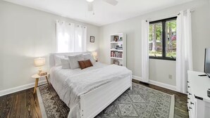 A bedroom with a bed, a nightstand, a bookshelf, a window with curtains, and a ceiling fan.