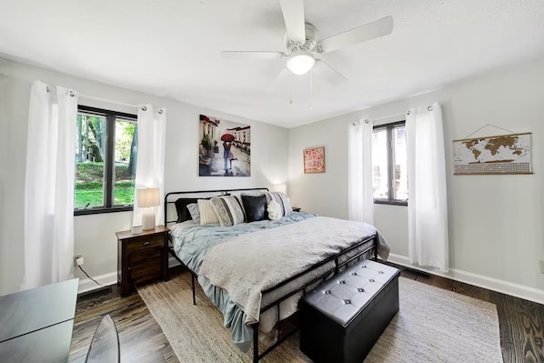 A bedroom with a bed, a nightstand, a desk, a chair, a window with curtains, a ceiling fan, and wall art.