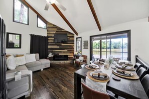 A modern living room with a fireplace, a dining area with a table set for six, and a large window offering a view of the outdoors.