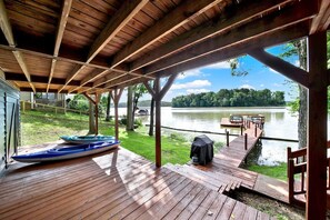 A covered deck with a view of a lake, a kayak, and a grill.