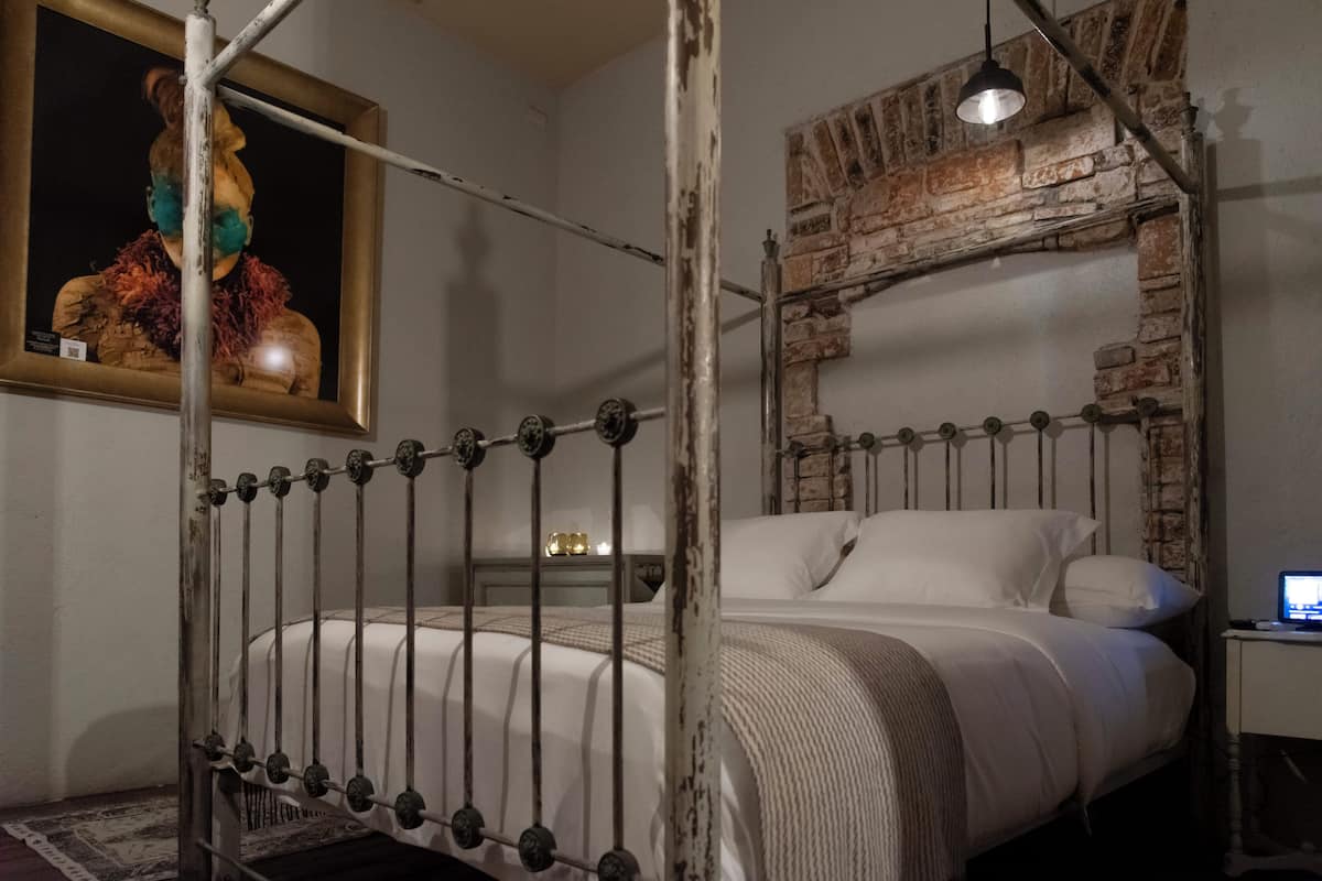 Ein Schlafzimmer mit einem Metallbettgestell, einer Ziegelwand, einem hölzernen Kopfteil, einem Nachttisch mit Lampe und einem Bild an der Wand.