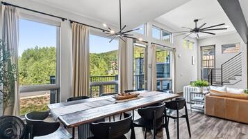 A modern dining area with a wooden table and chairs, large windows with curtains, and a view of a mountainous landscape.