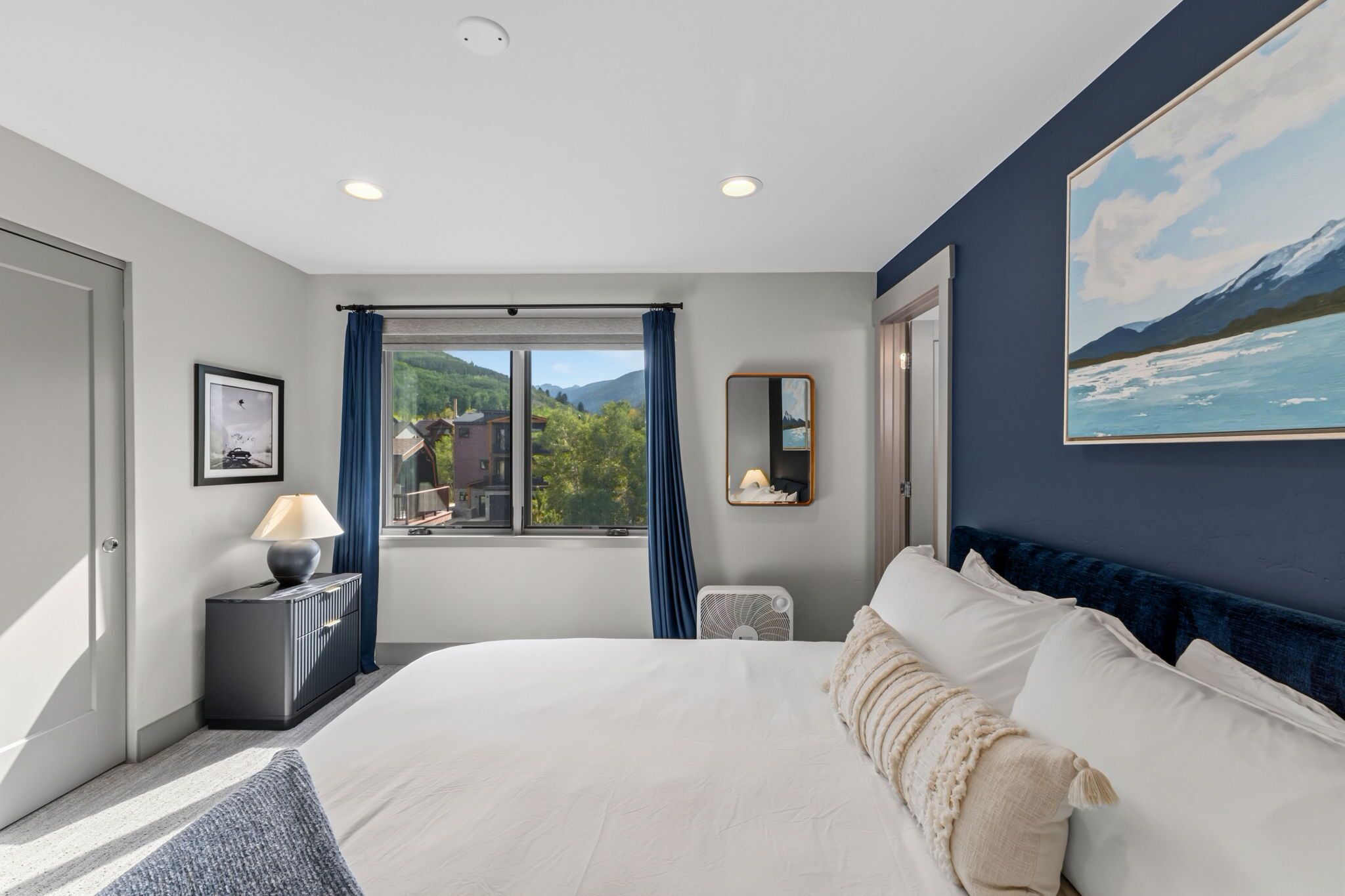 Een moderne hotelkamer met een groot bed, een nachtkastje met een lamp, een raam met uitzicht op groen en bergen, en een schilderij aan de muur.