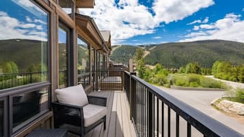 A balcony with a view of a forested mountain range, furnished with wicker chairs and a table.