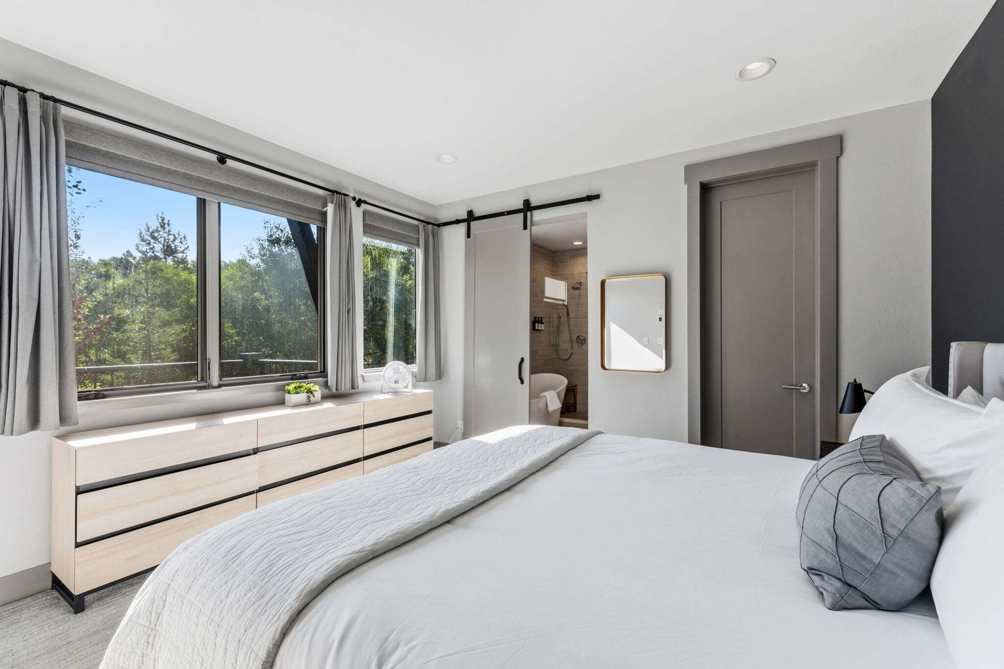 A modern bedroom with a large bed, a walk-in closet, a bathroom, and a view of trees through the window.