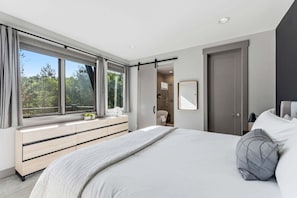 Ein modernes Schlafzimmer mit einem großen Bett, einem begehbaren Kleiderschrank, einem Badezimmer und Blick auf Bäume durch das Fenster.