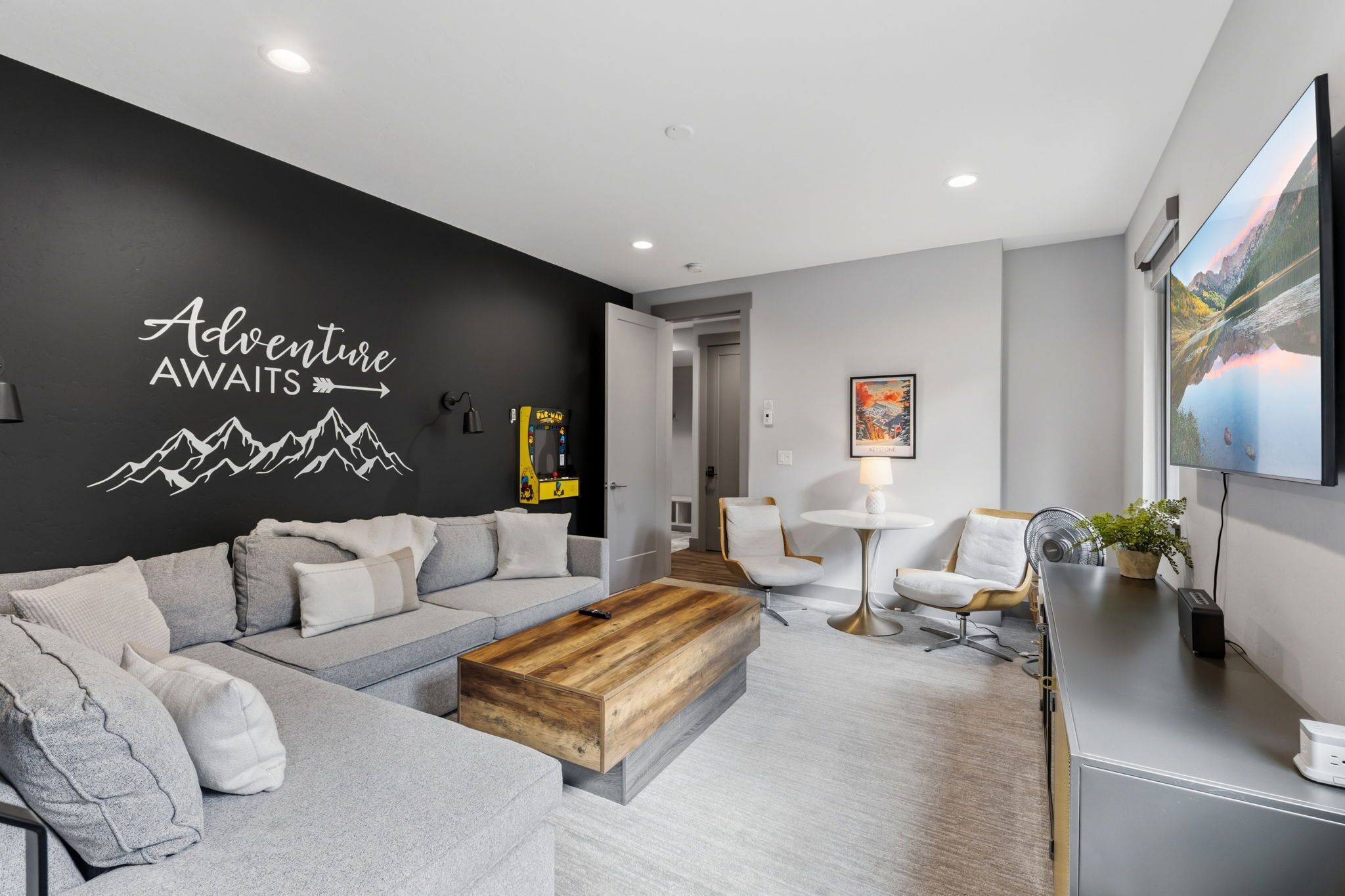 Een moderne woonkamer met een zwarte muur waarop een bergdesign en de tekst 'Adventure Awaits' staan. Er is een grijze bank, een houten bijzettafeltje en een klein, wit tafeltje met een lamp.
