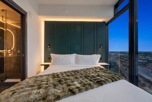 Ein modernes Hotelzimmer mit einem großen Bett, Blick auf die Stadt und einer Glastür zum Badezimmer.