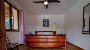 Quarto com cama de madeira, ventilador de teto, janela com cortinas e um quadro na parede.