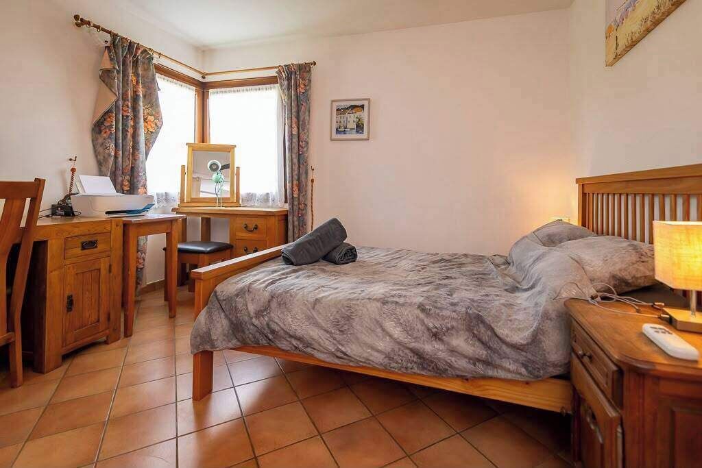 Une chambre à coucher avec un lit, un bureau, une chaise, une table de chevet et une fenêtre avec des rideaux.