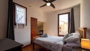 Quarto com cama, escrivaninha, ventilador de teto e janela com cortinas.