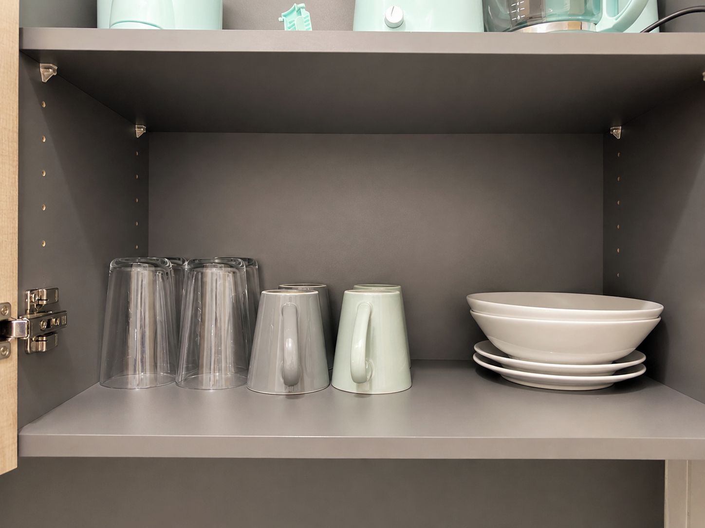 A kitchen shelf with clear glasses, a white pitcher, and stacked white plates.