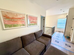 Ein Wohnzimmer mit einem braunen Sofa, zwei abstrakten Bildern, einer Treppe und einem Fenster mit Jalousien.