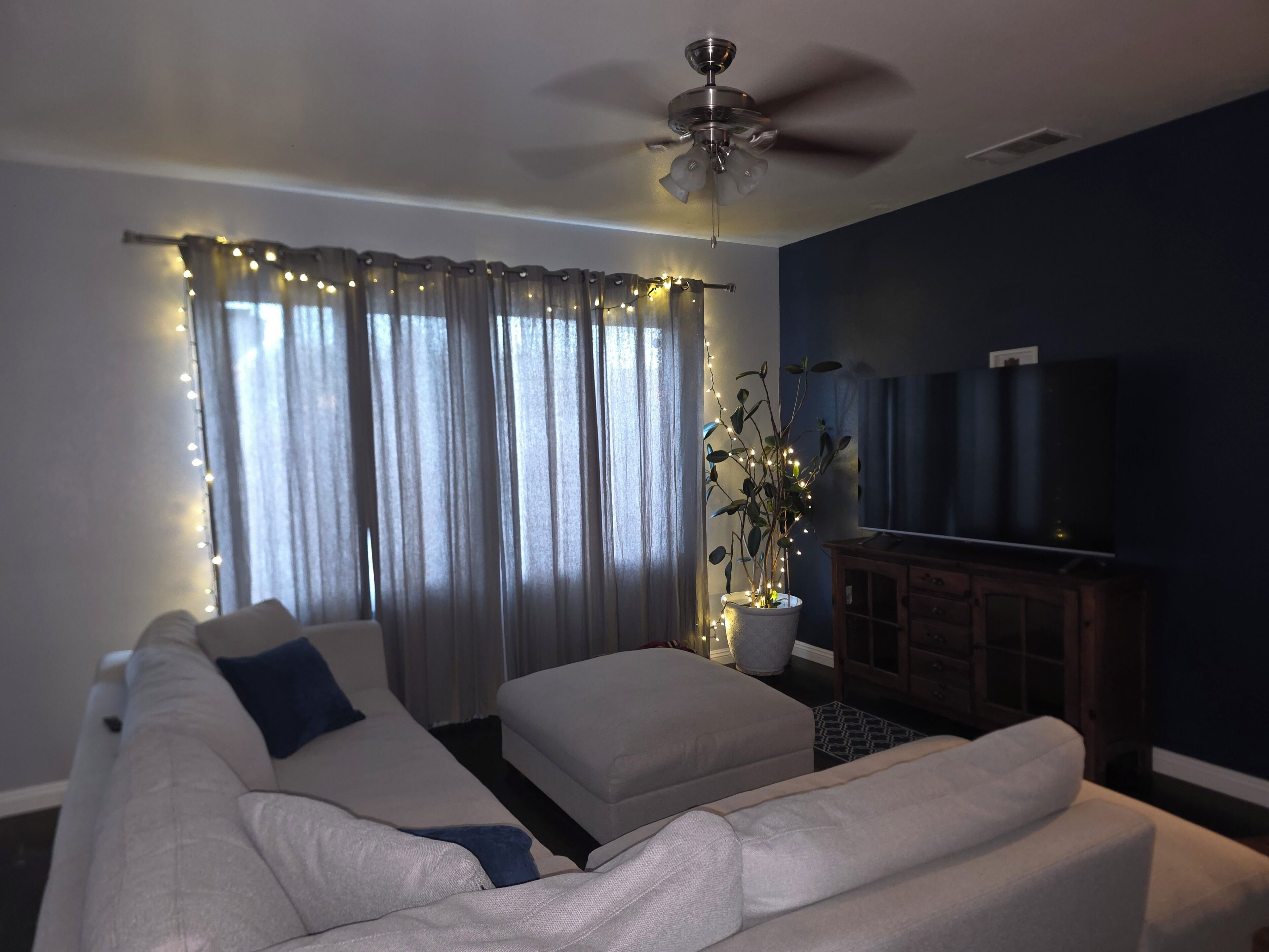 Una sala de estar con ventilador de techo, sofá, televisor y una planta con luces.