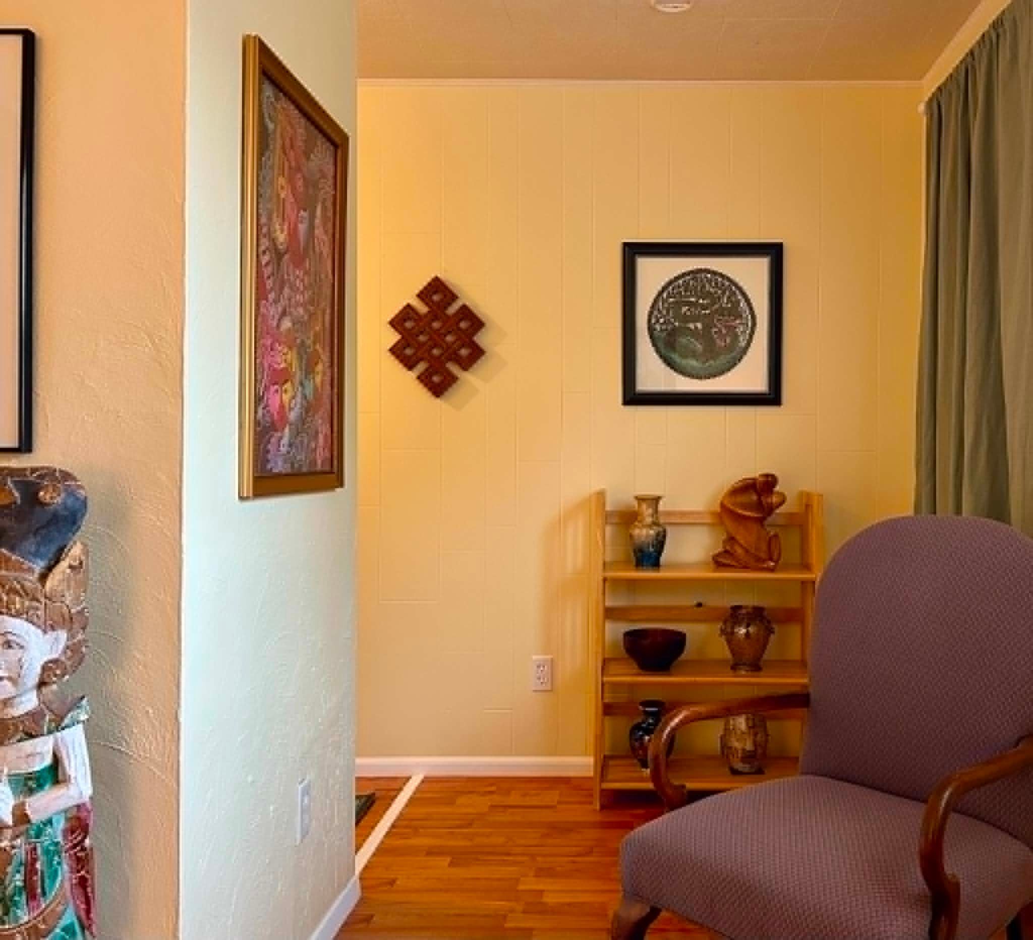 A living room with a wooden floor, a chair, a shelf with decorative items, and two framed pictures on the wall.