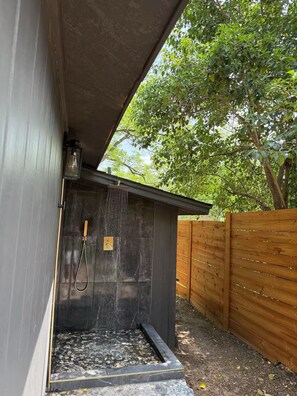A stone shower enclosure with a tiled floor, a wooden fence, and a tree in the background.