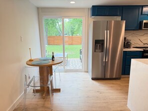 A modern kitchen with a stainless steel refrigerator, a dining area with a table and chairs, and a sliding glass door leading to a backyard.