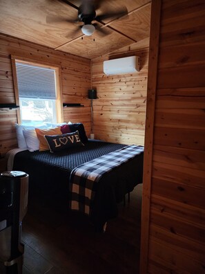Un dormitorio de cabaña de madera con una cama, un ventilador de techo, una ventana con persianas y un calentador fijado a la pared.