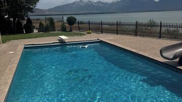 A rectangular swimming pool with clear blue water, surrounded by a paved deck. In the background, there is a mountain range and a body of water.