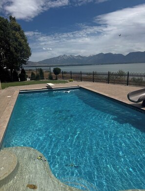 Una piscina rectangular con agua azul cristalina, rodeada por una solana empedrada. Al fondo, se divisan montañas y una masa de agua.