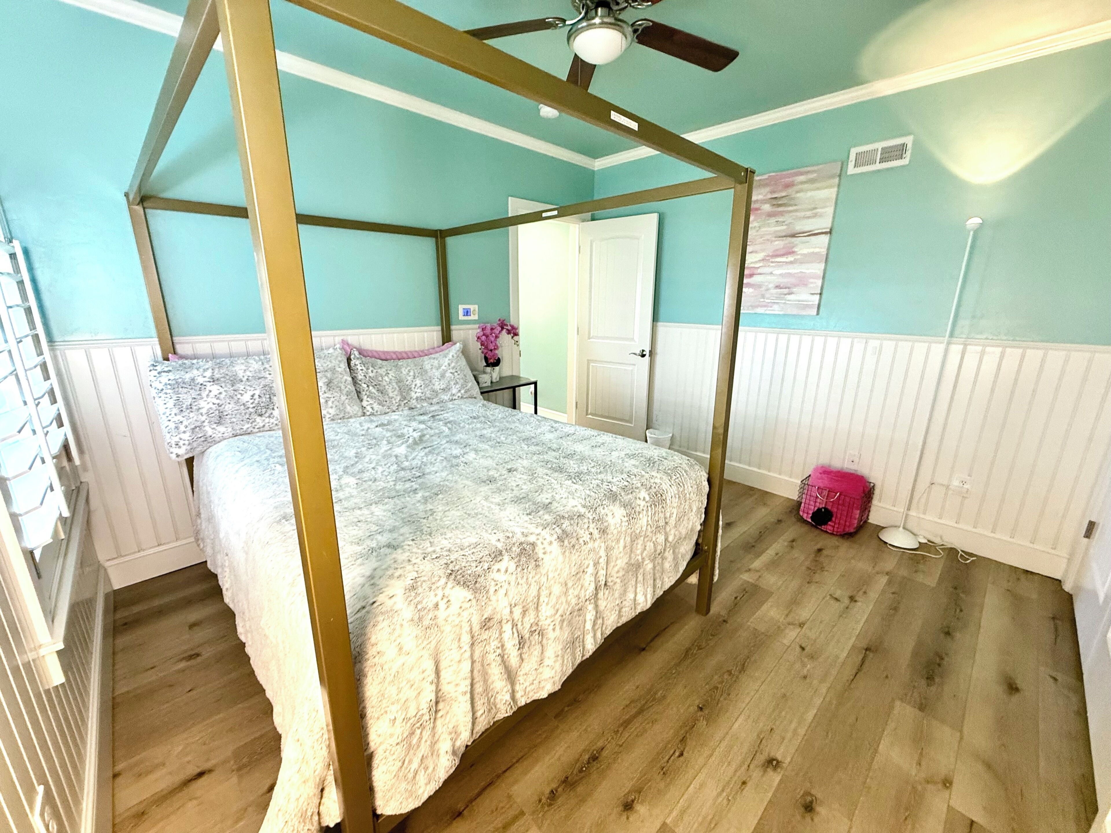 A four-poster bed with a canopy, a wooden floor, a white door, and a wall-mounted fan.