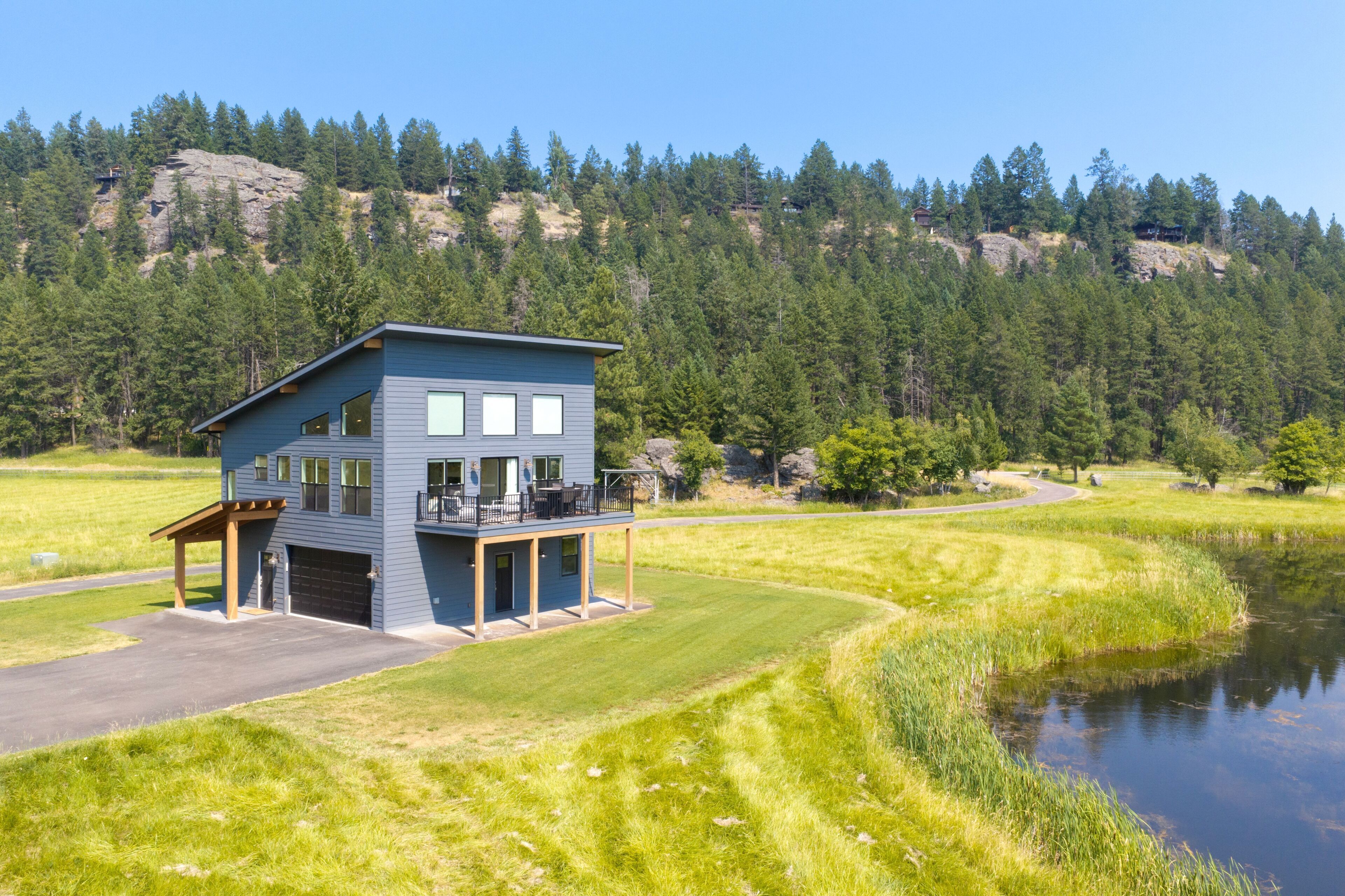 A modern two-story house with a garage and a deck, surrounded by a lake and forest.