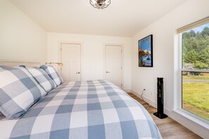 A bedroom with a checkered bedspread, two doors, a framed picture of Glacier National Park, and a window with a view of greenery.