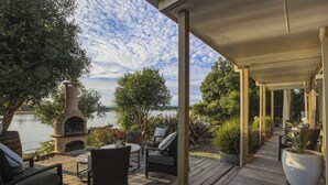 An outdoor deck with a stone fireplace, wicker furniture, and a view of a lake. - Estuary Edge in Mapua a luxurious getaway (Māpua)