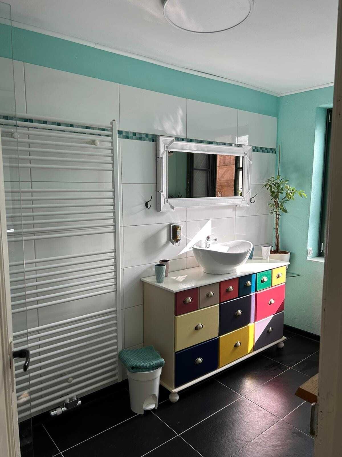 A bathroom with a white sink, a mirror, a colorful cabinet, a window, and a plant.