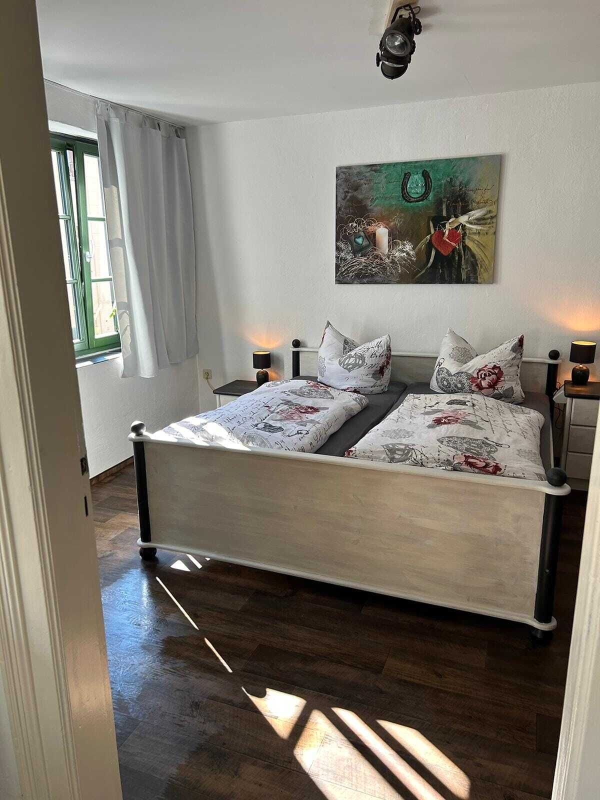 Ein Schlafzimmer mit einem Bett, zwei Nachttischlampen, einem Bild an der Wand und einem Fenster mit Vorhängen.