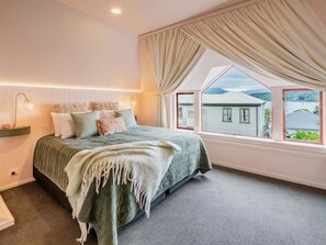 Ein Schlafzimmer mit einem großen Bett, einer Nachttischlampe, einem Fenster mit Vorhängen und Blick auf ein Haus.