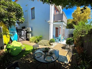 Une terrasse avec une table dressée pour deux personnes, entourée de verdure et bordée d’un bâtiment blanc doté d’un balcon.