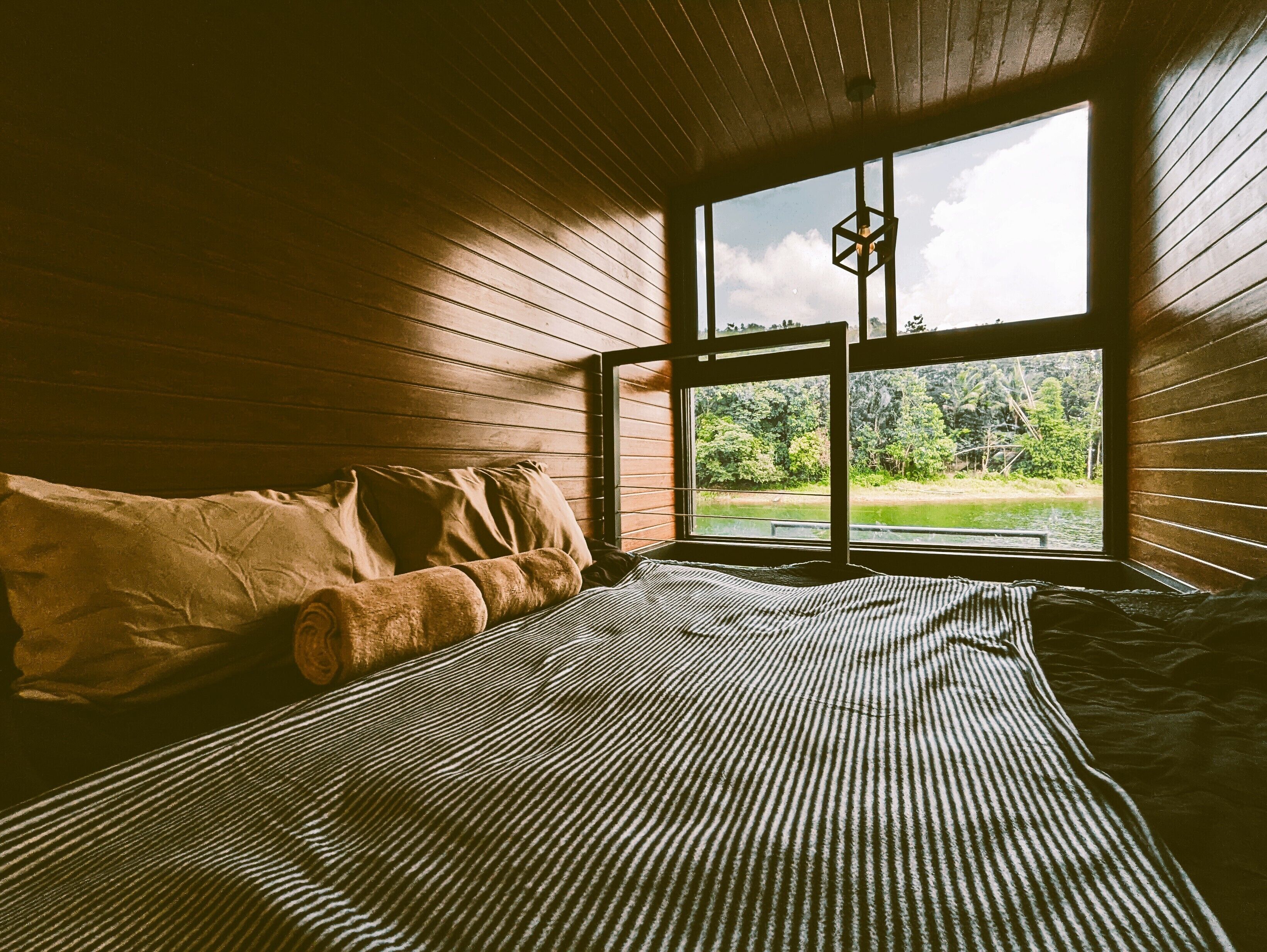 A room with a bed, pillows, and a window with a view of a forest and water.