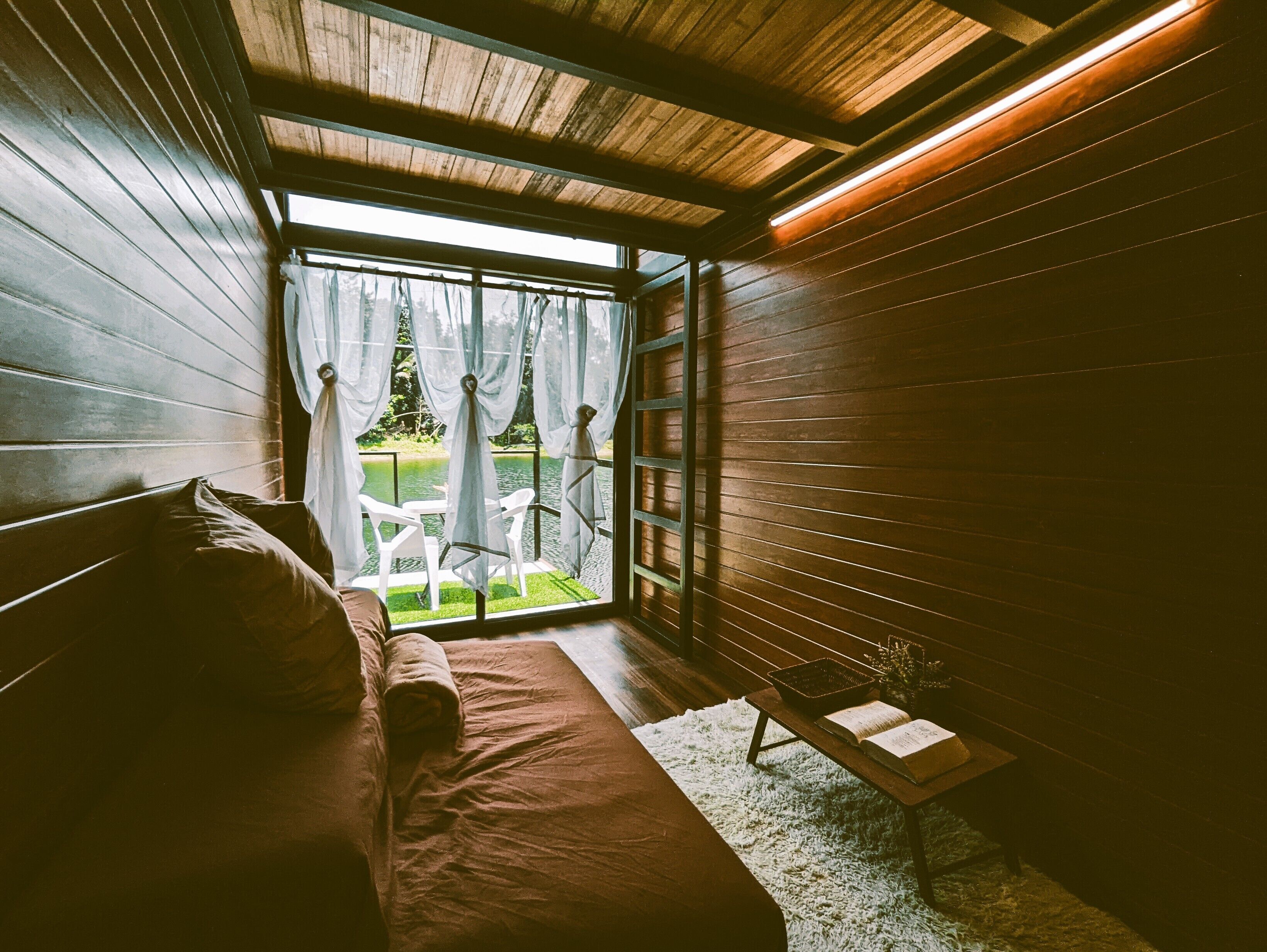 Ein gemütliches Zimmer mit Holzbalkendecke, einer Bank, einem kleinen Tisch mit einem Buch und Blick auf einen Garten durch ein Fenster.