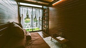 A cozy room with a wooden ceiling, a bench, a small table with a book, and a view of a garden through a window.