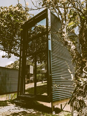 A modern, minimalist structure with a glass door, surrounded by lush greenery and a clear sky.