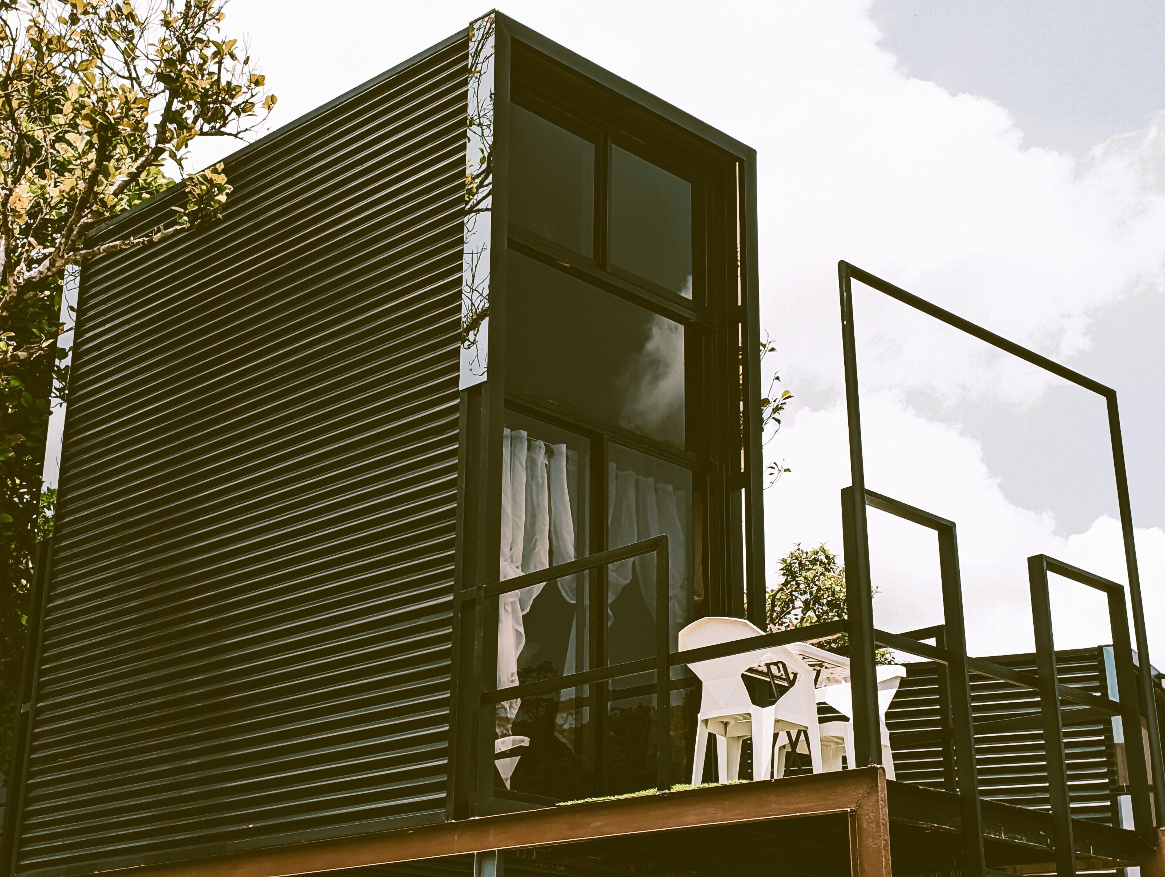Un bâtiment moderne doté d’un toit en pente, de grandes fenêtres et d’un balcon avec des chaises en plastique.