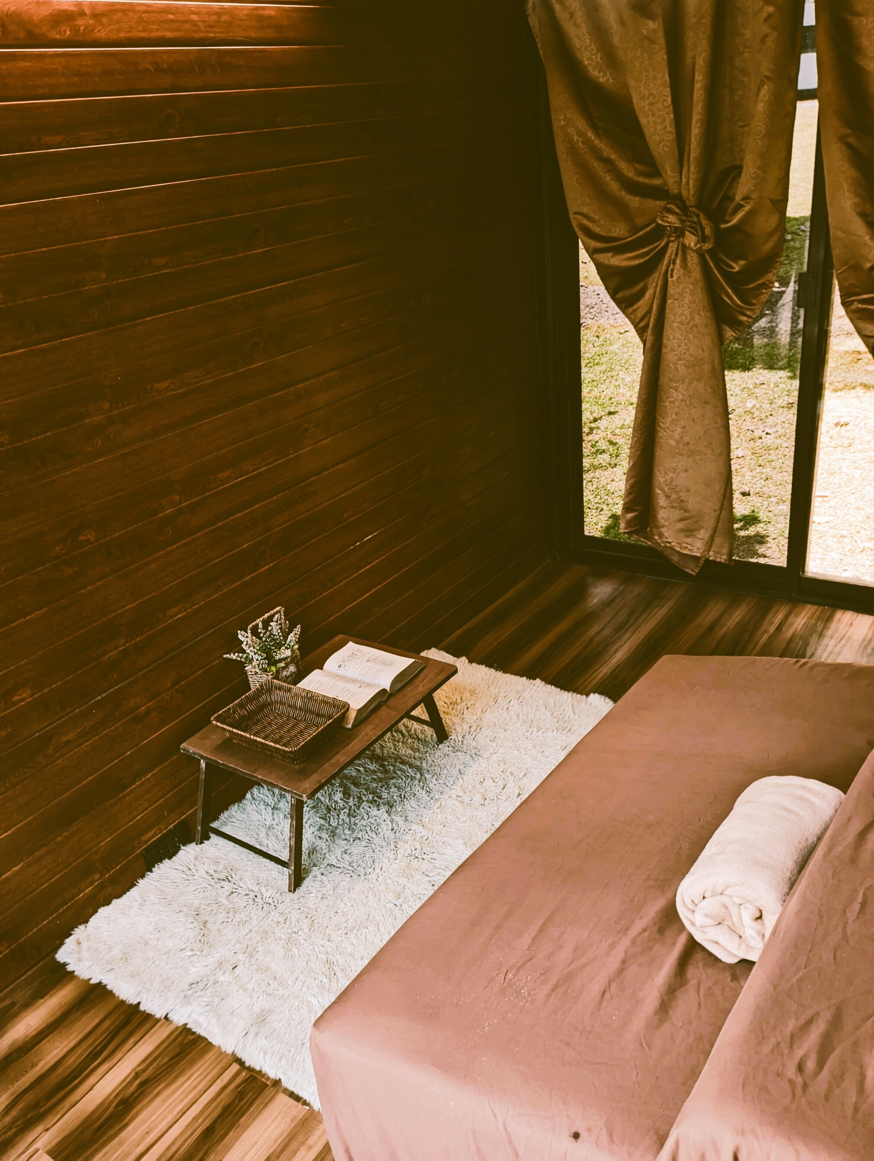 Een slaapkamer met een houten vloer, een wit tapijt, een bed met een roze sprei, een klein houten tafeltje met een mand en een boek, en een raam met gordijnen.