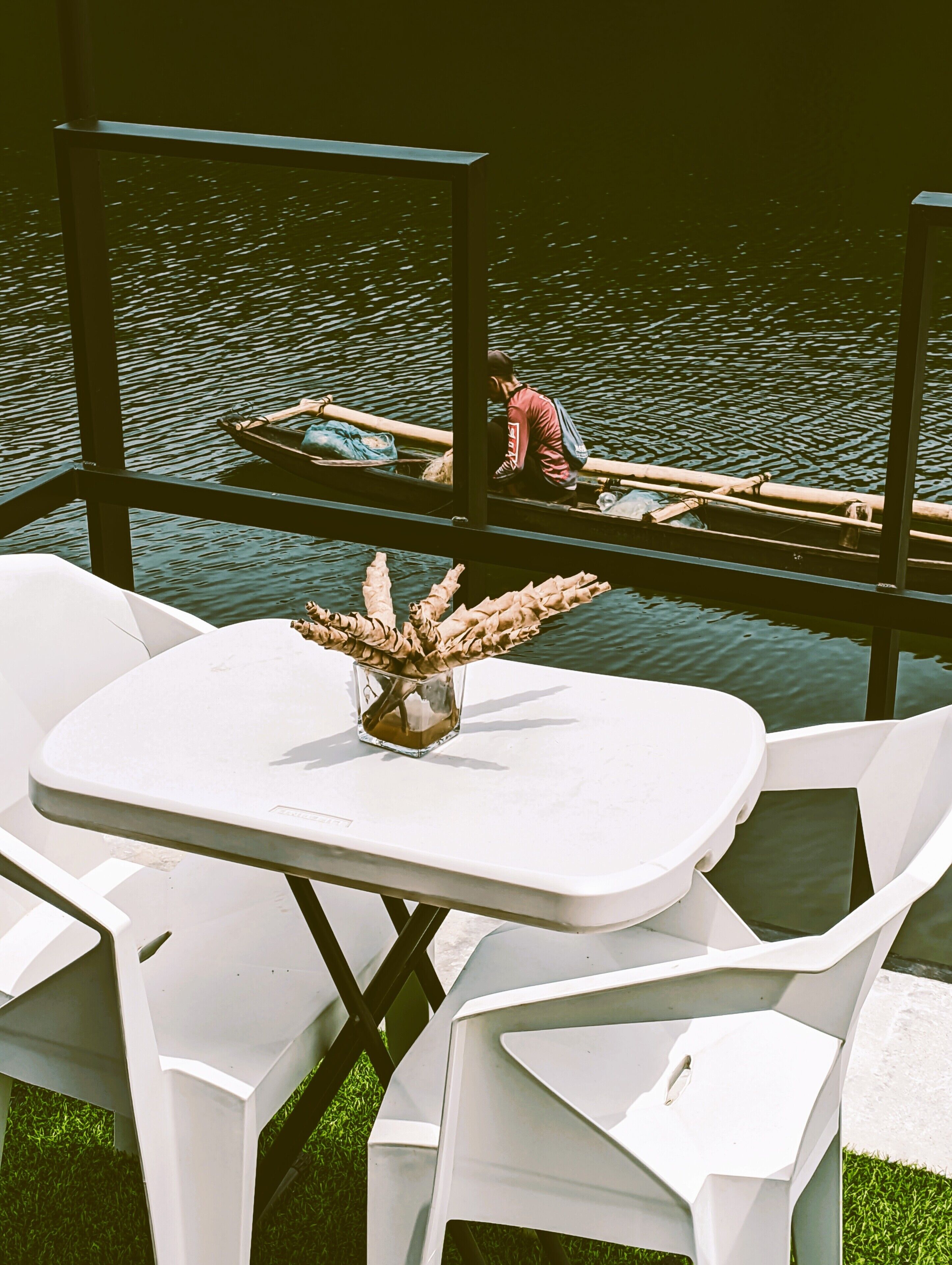 Een tafel met stoelen en een vaas droogbloemen, met uitzicht op een rustig waterlichaam waar iemand een boot roeit.
